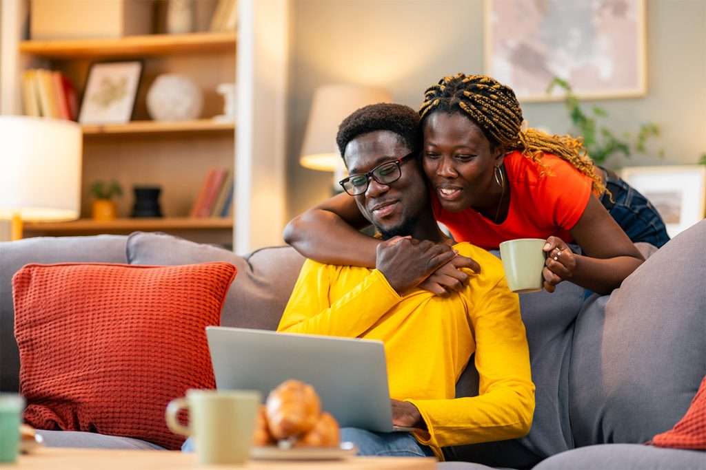 Couple sitting on couch smiling and looking at a laptop.
