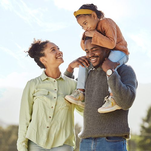 Photo of a mom and dad with their child on the father's shoulders. Outside with a blue sky, smiling.